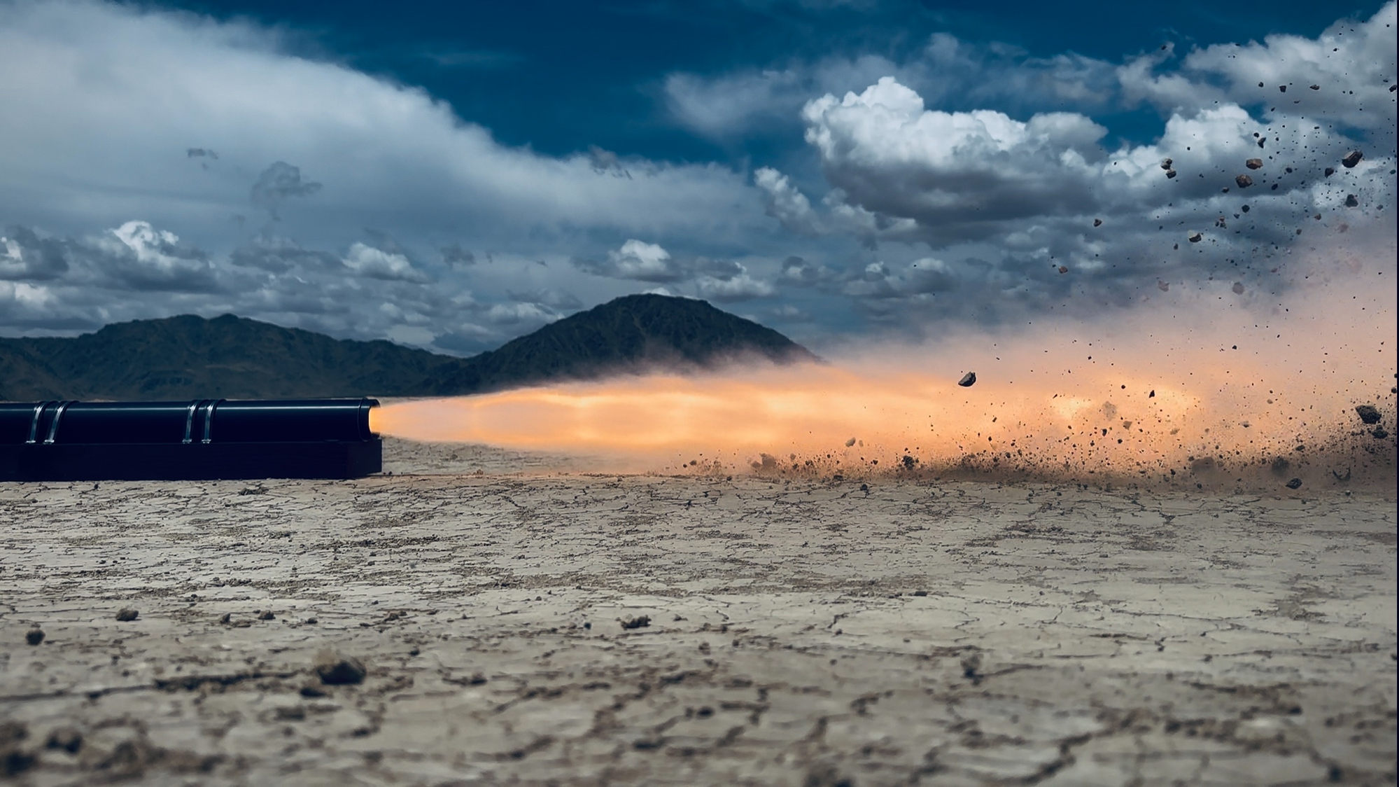 Solid rocket motor hotfire on a Mojave dry lake bed, with a long orange plume cutting across the cracked white playa under stormy blue sky.
